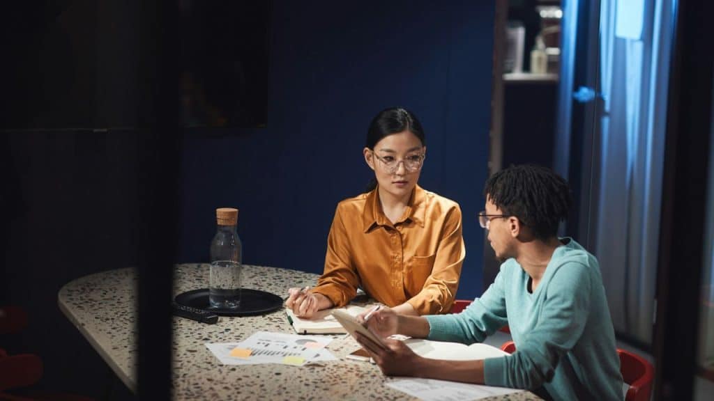 A woman in a gold shirt and a man in teal sit at a table with papers.