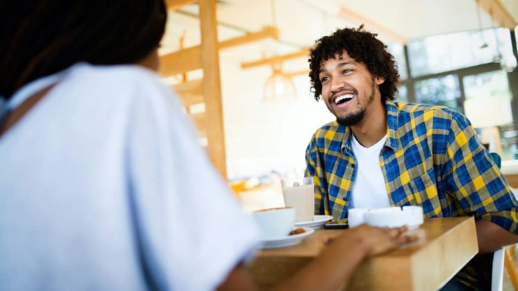 A man with curly hair and a plaid shirt laughs while sitting across from someone.