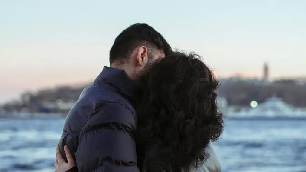 A couple embraces near the water at sunset.