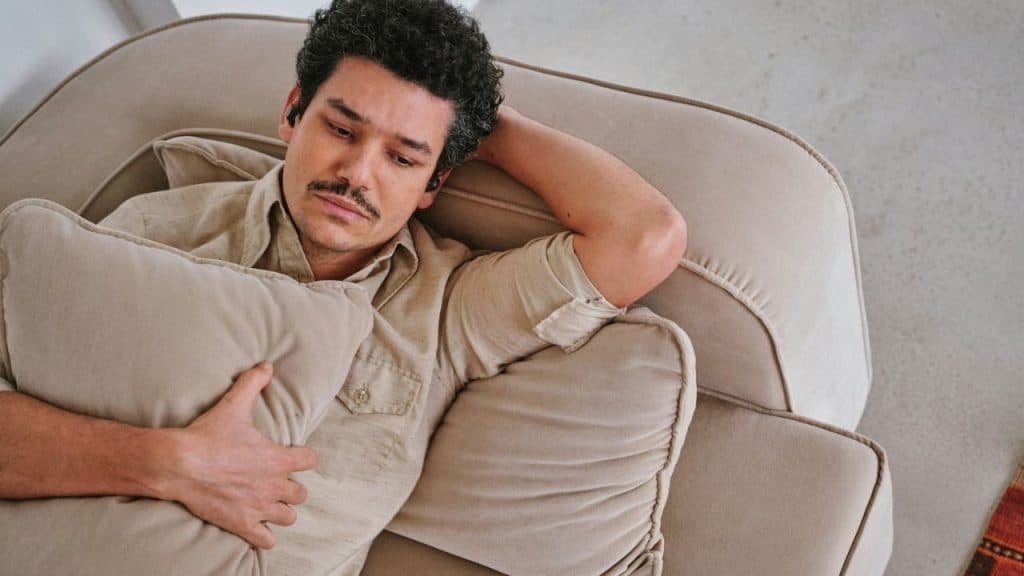 A man with curly hair and a mustache lies on a tan sofa, clutching a pillow.