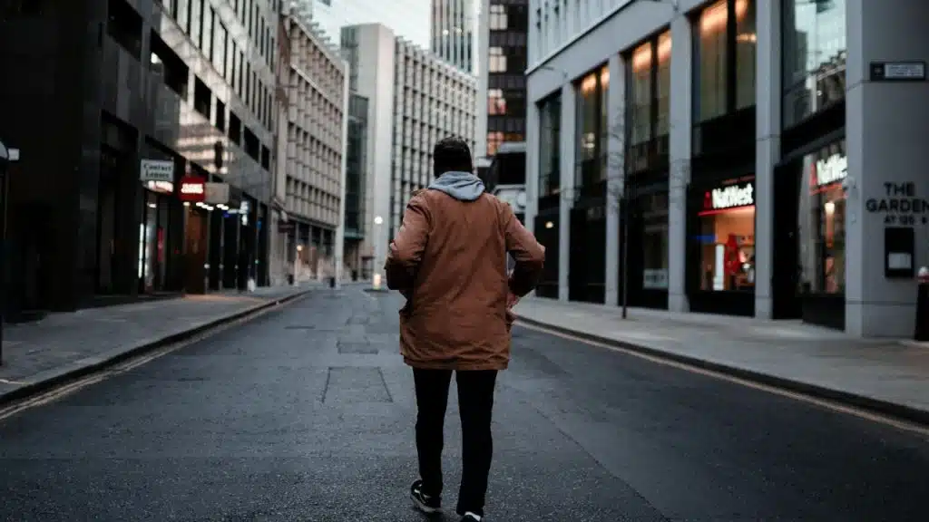A man in a brown jacket and grey hoodie walks away down a city street.