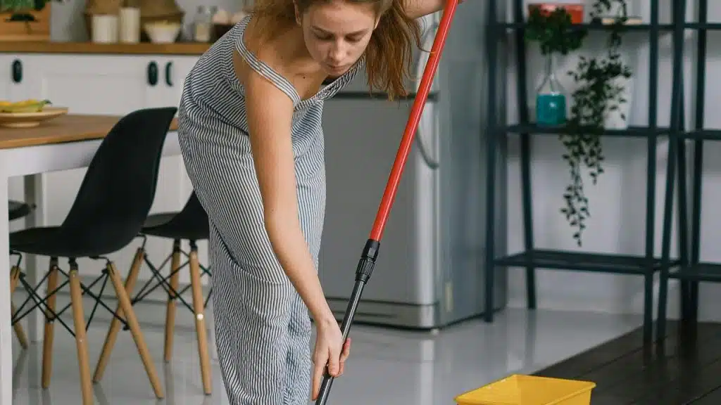 A woman mopping a kitchen floor with a red-handled mop and a yellow bucket nearby.