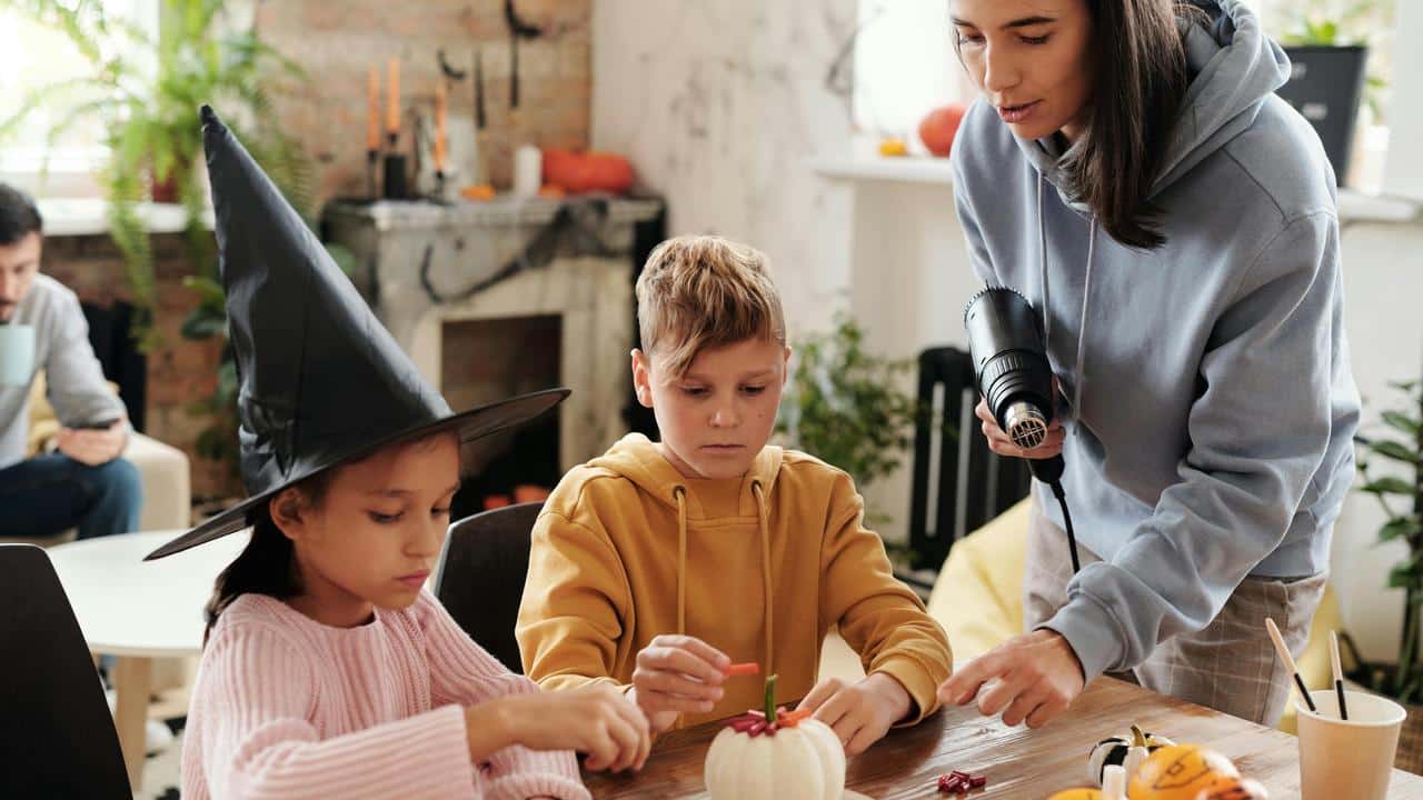 A woman helps two children decorate a pumpkin, with one child wearing a witch hat.