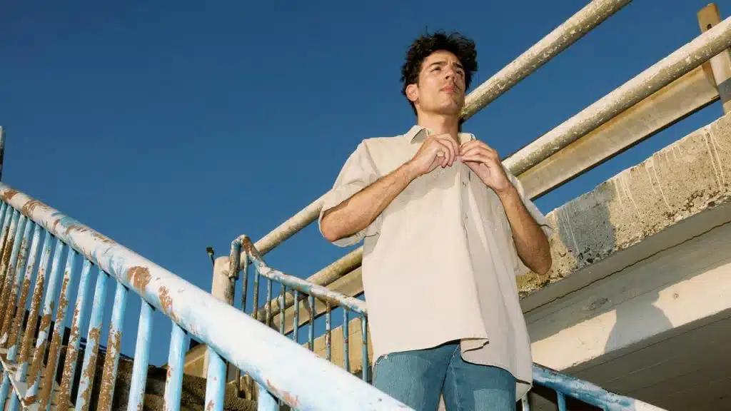 A man buttoning his shirt on an outdoor staircase under a clear blue sky.