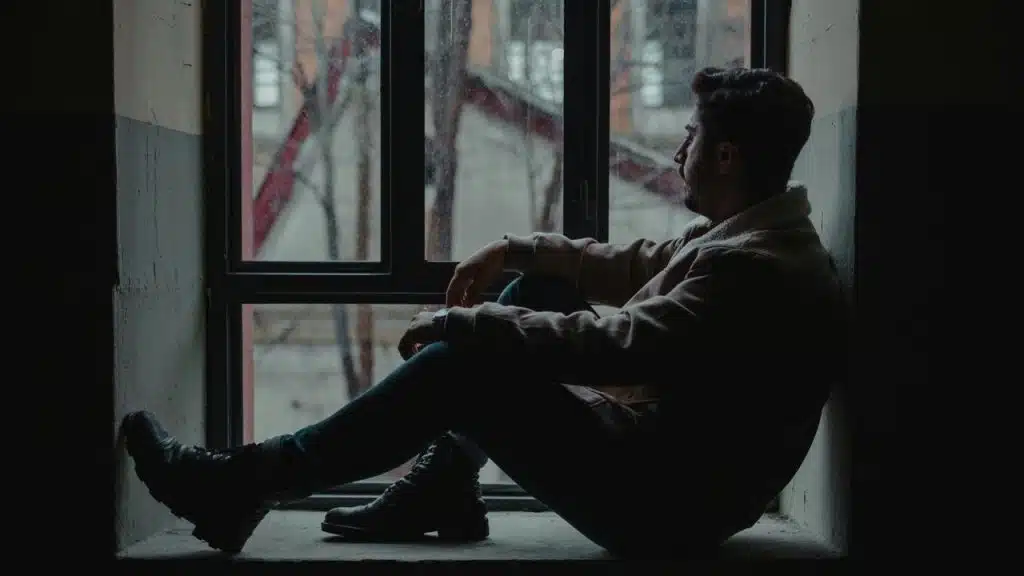 A man sitting on a windowsill indoors, gazing out through a dimly lit window.
