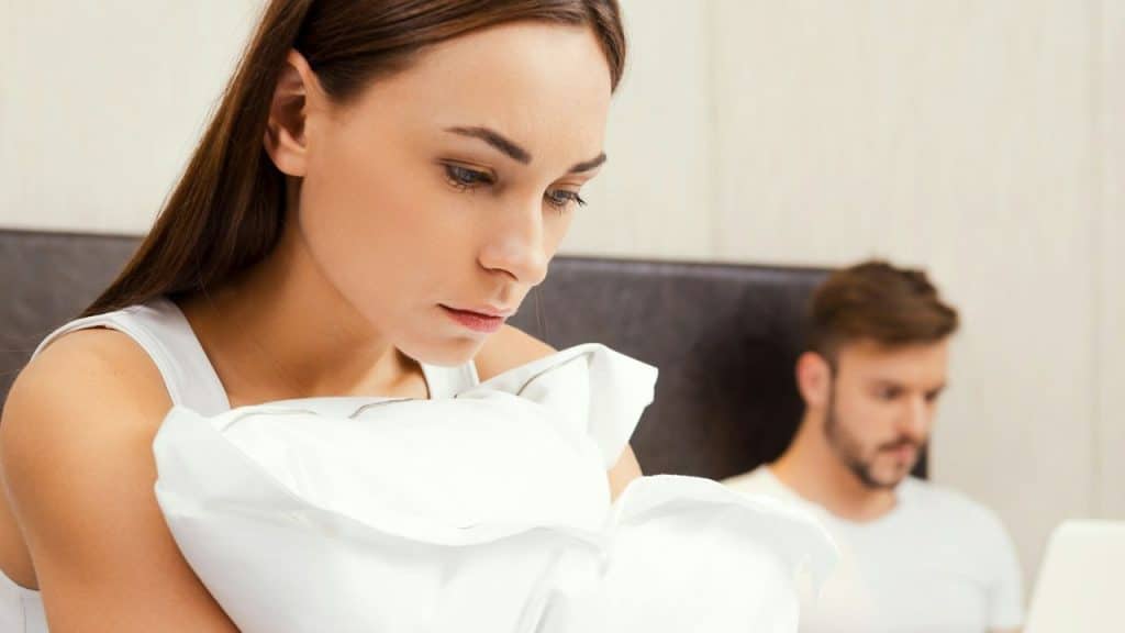 A woman in a white tank top looks down while a man sits blurred behind her.