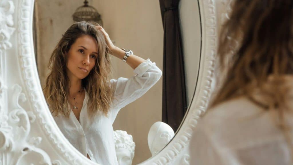 A woman in a white shirt looks at her reflection while touching her hair.