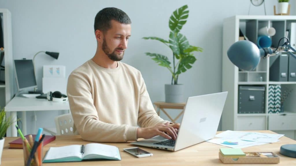 A bearded man types on a laptop at a wooden desk in a bright office.