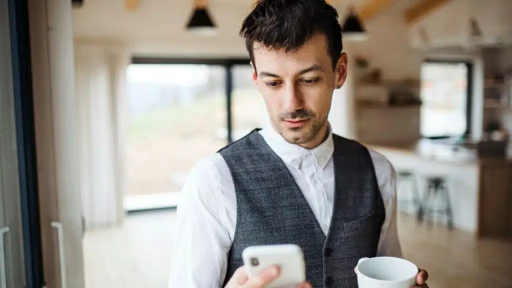 A man in a white shirt and gray vest looks down at his phone while holding a white cup.