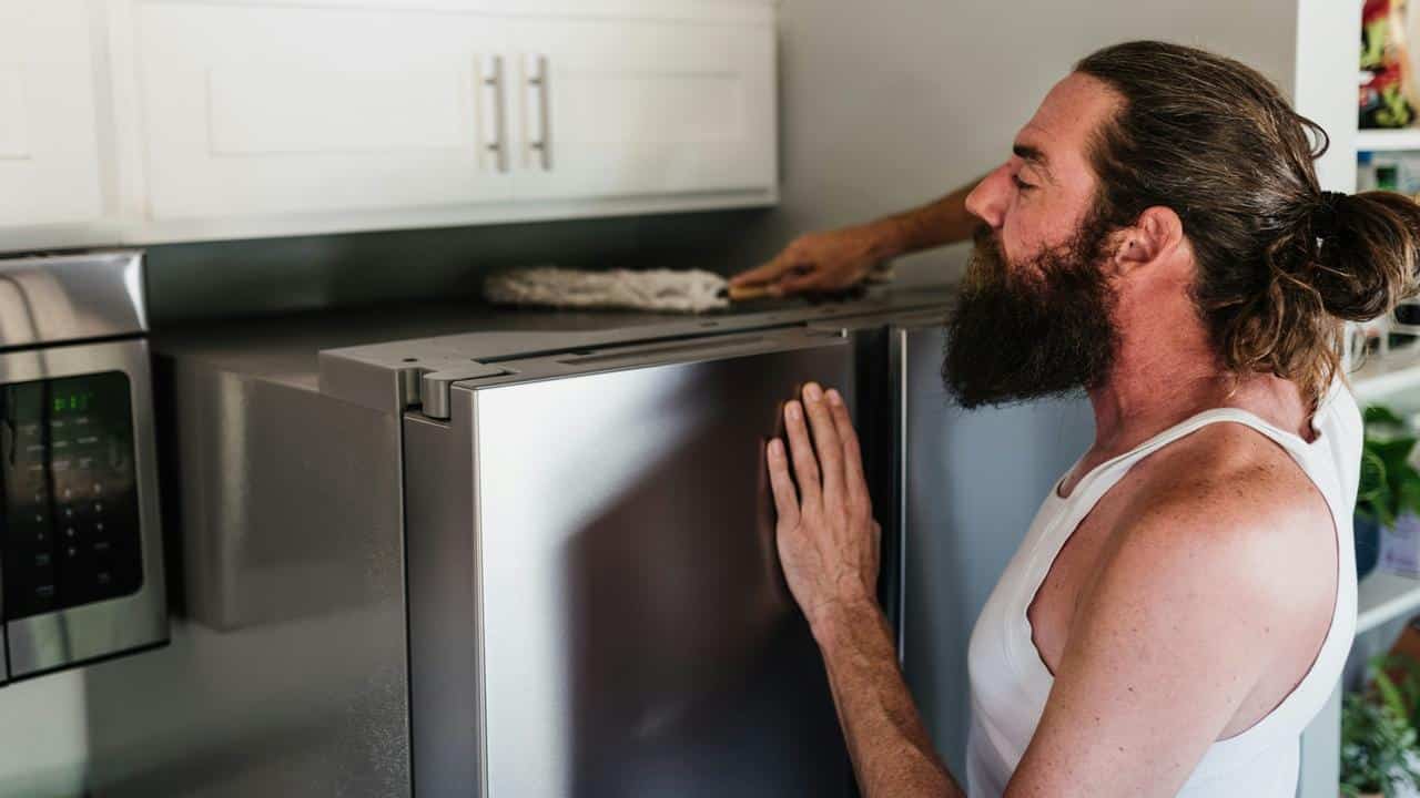 A man cleaning the top of a refrigerator in his kitchen.