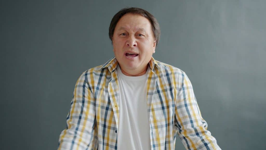 Middle-aged man in a plaid shirt speaking against a gray background.