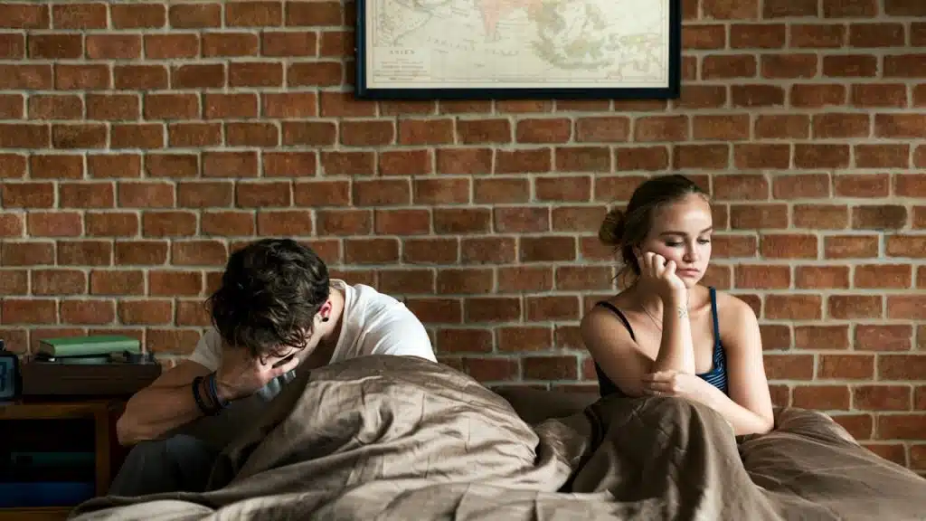 Man and woman sitting up in bed, looking sad and distant from each other.