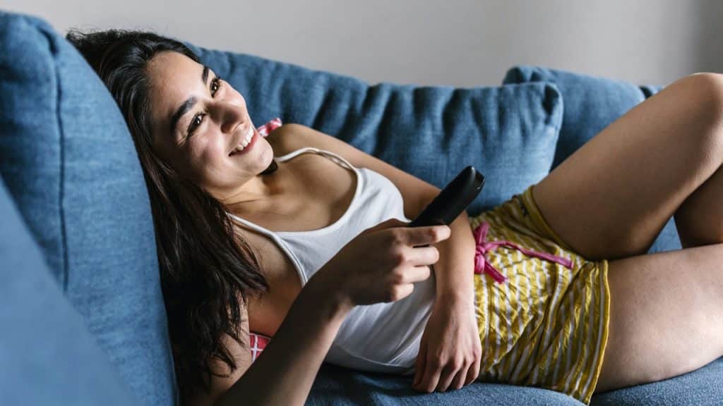 A young woman is relaxing on a blue couch, holding a remote control and smiling while watching TV.