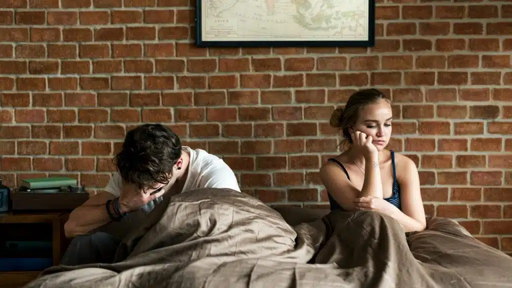 Man and woman sitting apart on a bed, looking upset after an argument, with a brick wall behind them.