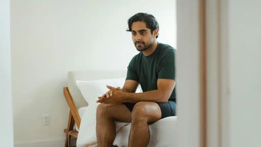 A man in a dark T-shirt and shorts sits on a bed with his hands clasped, looking away.