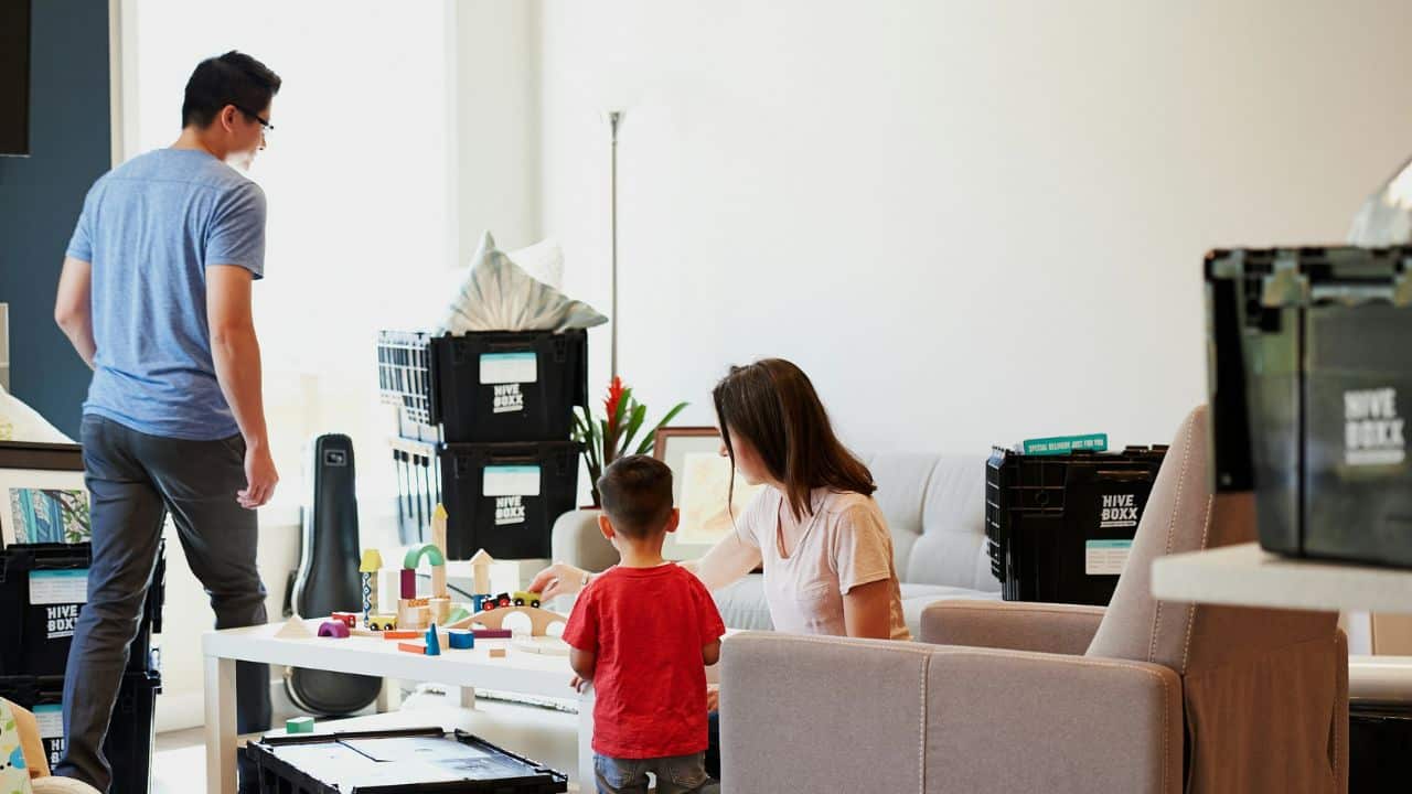 A family is in a bright living room surrounded by black moving bins, playing with wooden blocks.