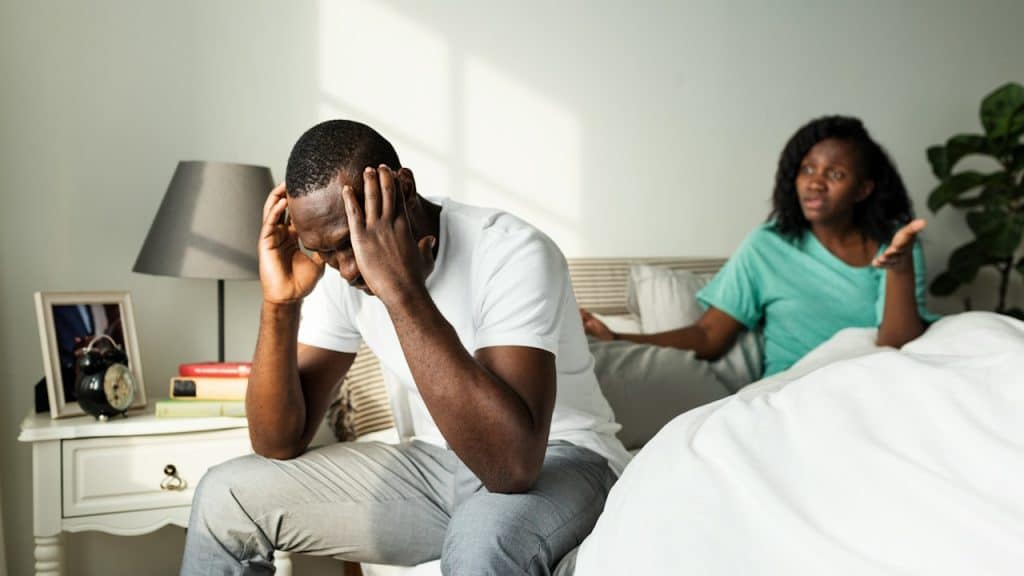A woman complaining to her husband while he is sitting next to her holding his head.