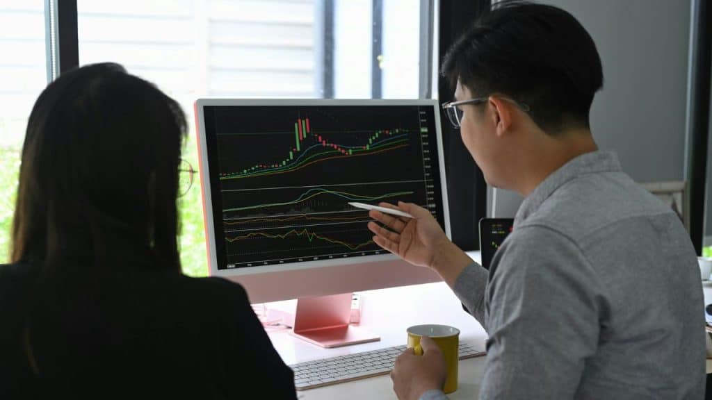 Two people look at a computer screen displaying complex stock or trading charts.