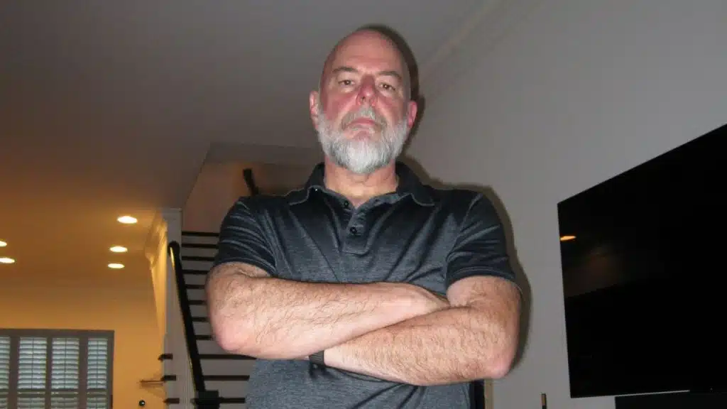 Bald man with a white beard wearing a black polo shirt, standing with arms crossed.