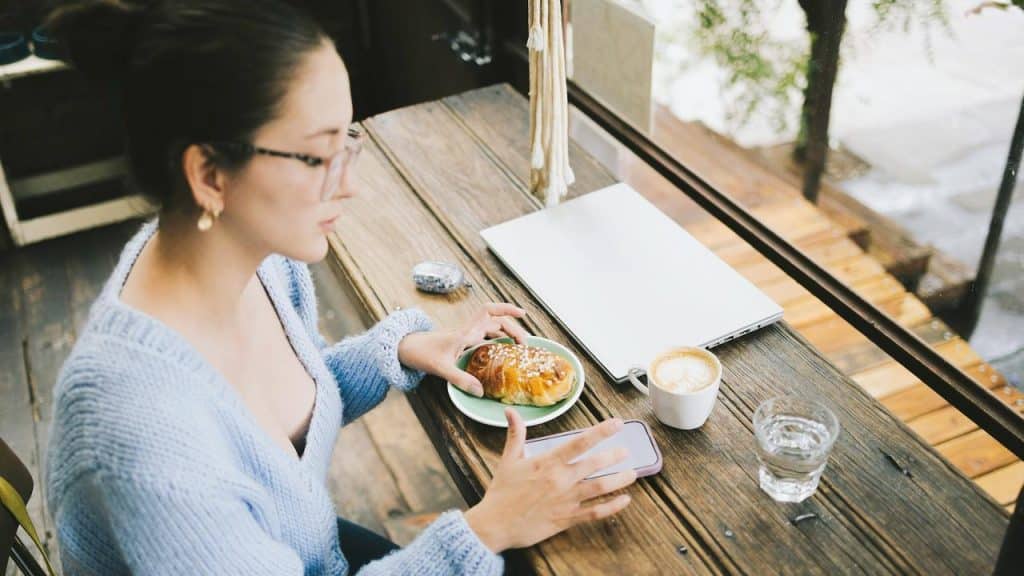 A woman in glasses sits at a café table with a croissant, coffee, and a laptop.