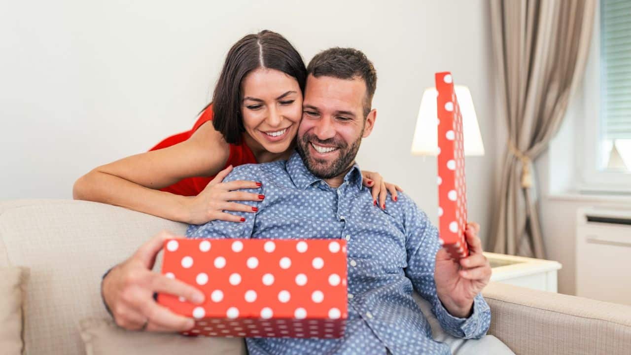 Smiling couple opening a red and white polka dot gift box together on a couch.