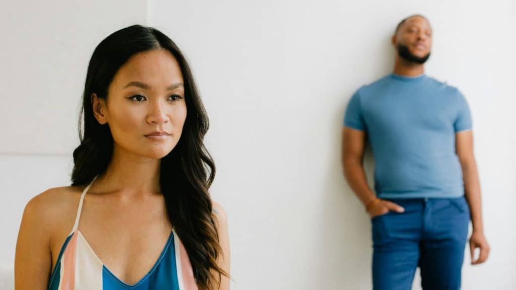 A woman in a striped top looks away while a man leans against a wall in the background.