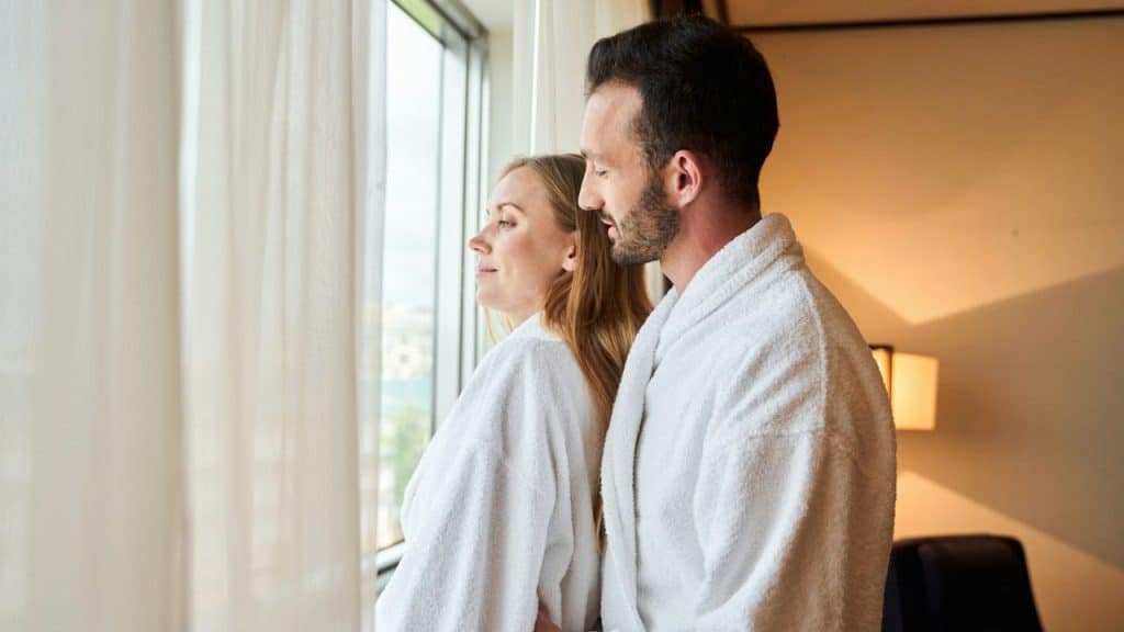Couple in white robes stands together looking out a window covered by sheer curtains.