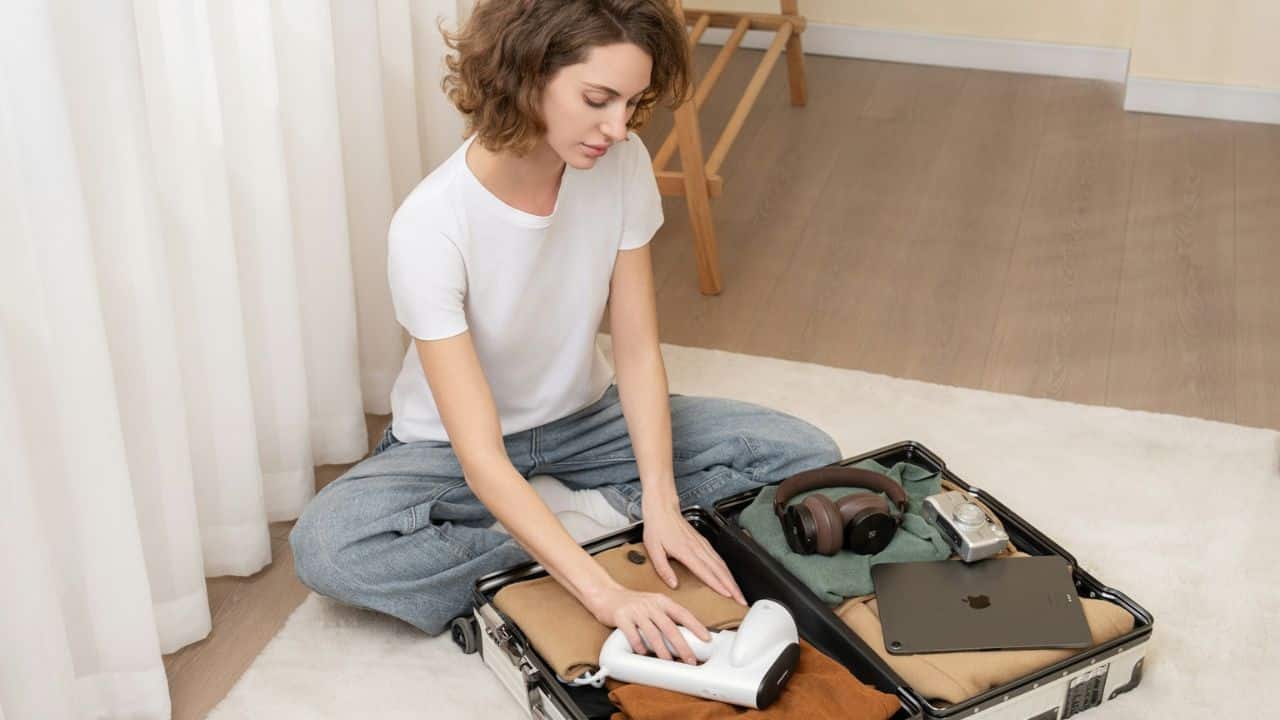 Woman sitting cross-legged on the floor, packing clothes and electronics into a suitcase.
