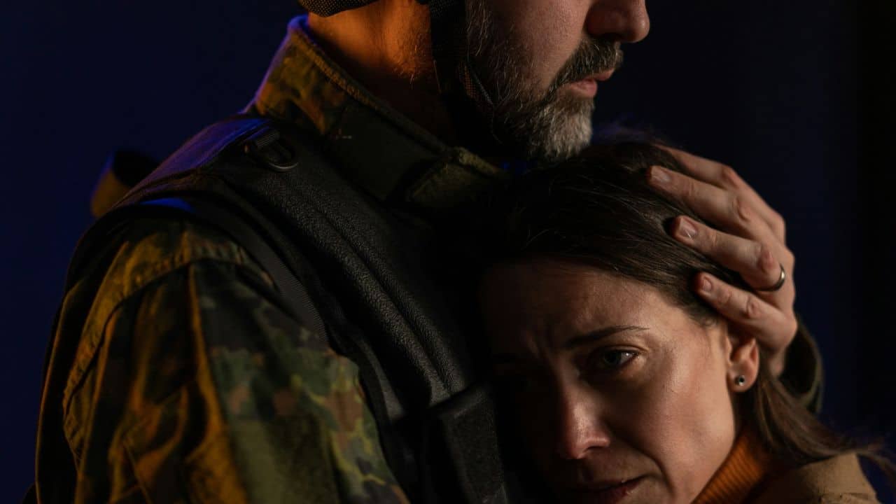 Woman with worried expression being embraced by a man in military-style gear.