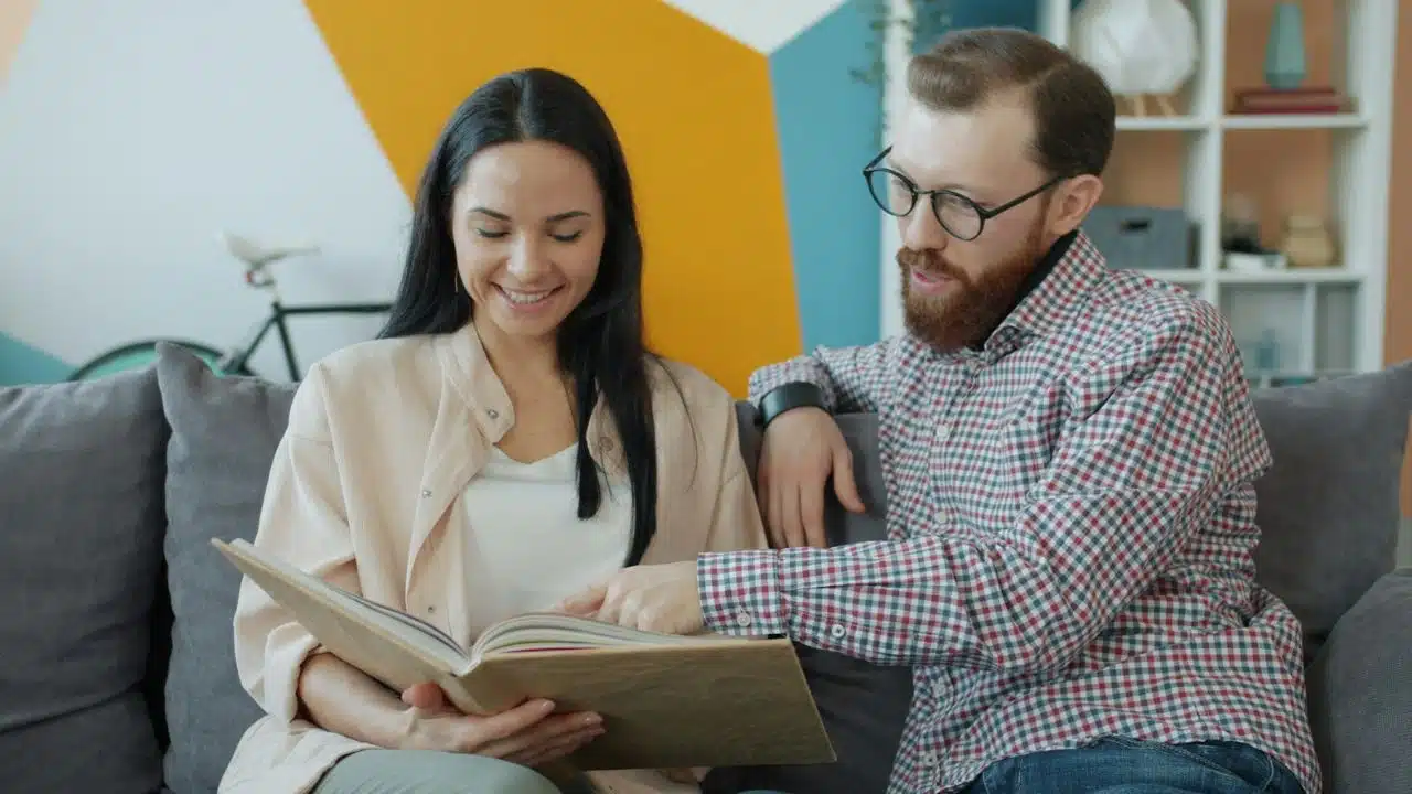A smiling couple sits on a couch looking at an open photo album together.