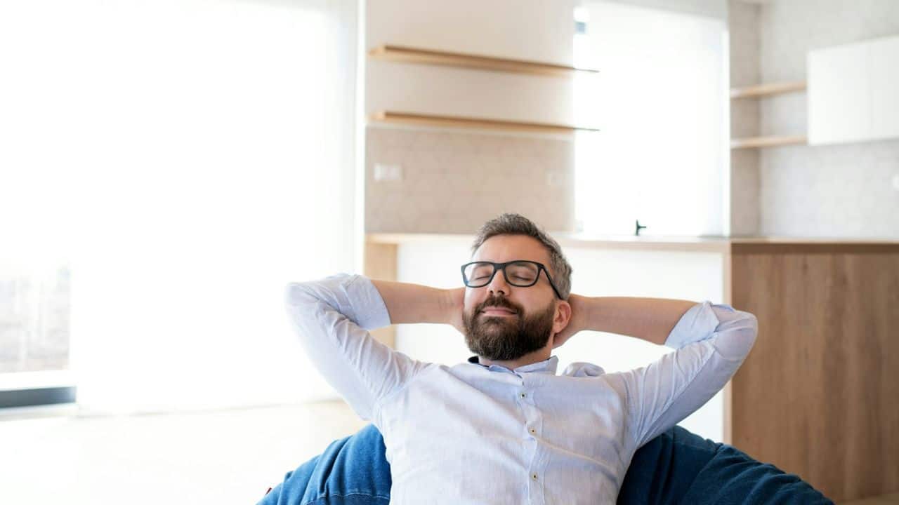 A bearded man wearing glasses is relaxing with hands behind his head indoors.