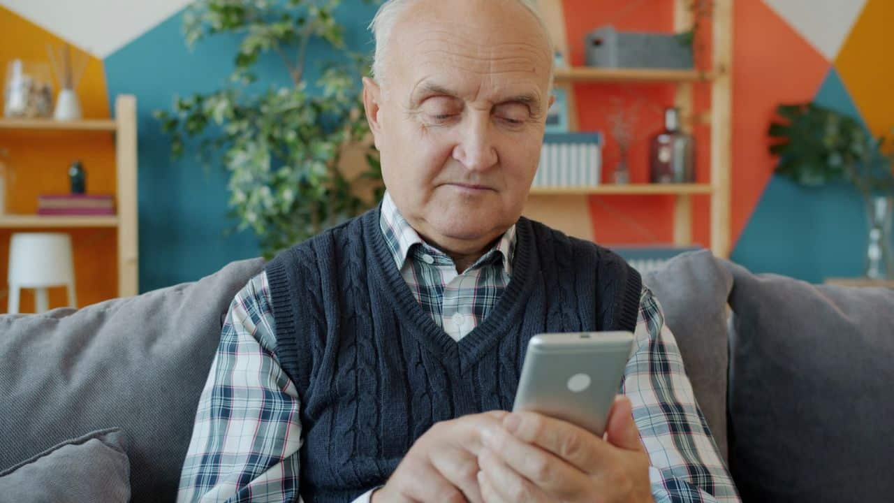 An older man sits on a couch, looking at his smartphone indoors.