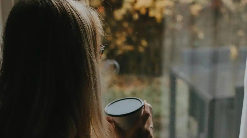 A woman with long hair holds a cup and looks out the window.
