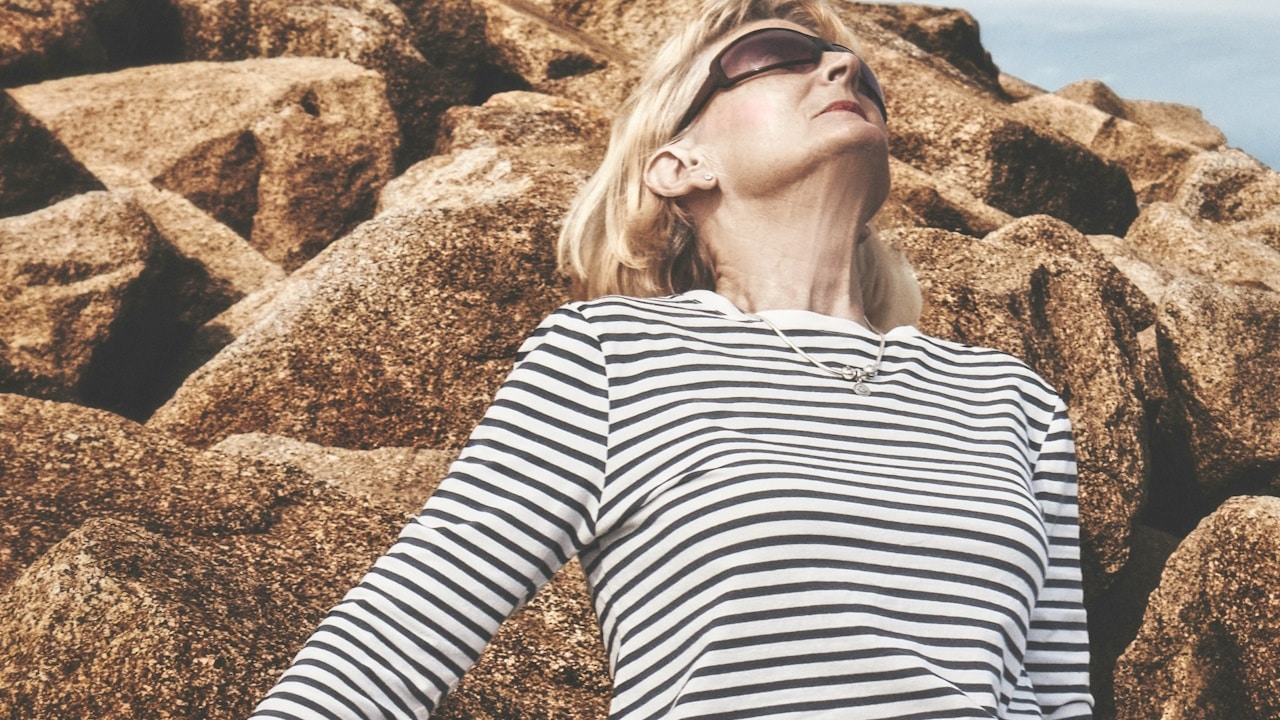 An old modern woman leaning against rocks on a beach.