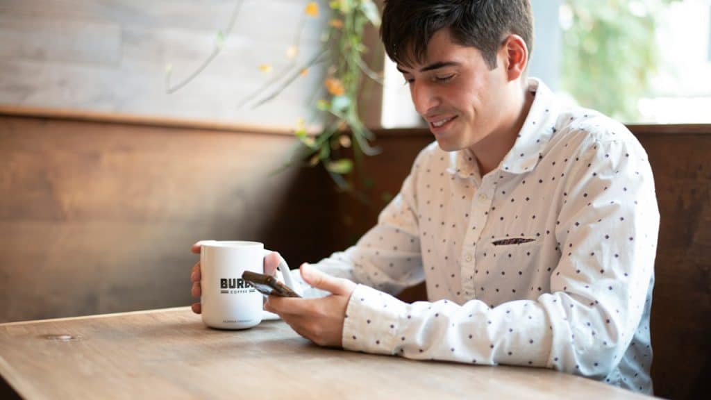 A man smiling while having coffee and looking at his phone as he sits at a table.