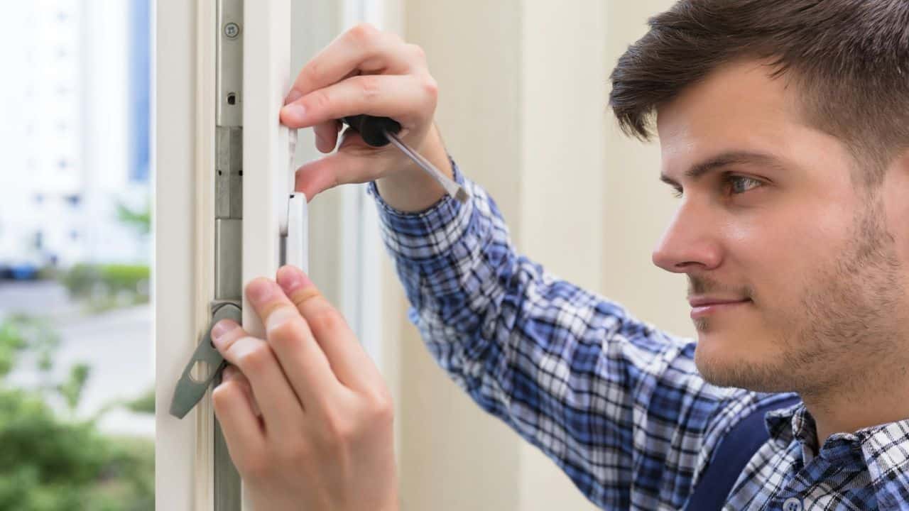 A young man in a plaid shirt is using a small screwdriver to work on a white window frame or lock mechanism.