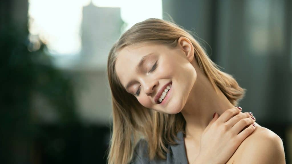A young smiling woman touching her neck and collarbone.