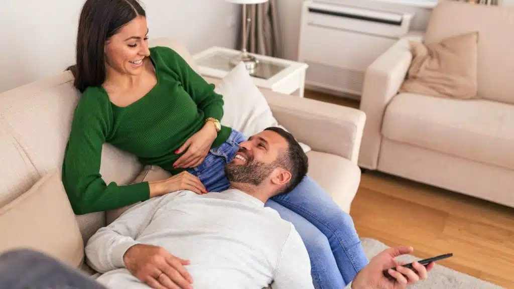A smiling woman is lying with her head in a man's lap on a light-colored couch.