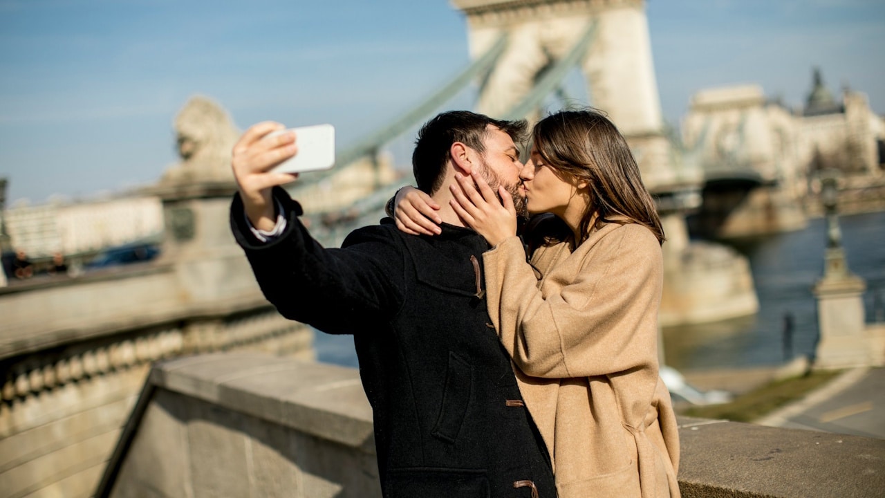 A couple is kissing and taking a selfie near a bridge landmark on a sunny day.