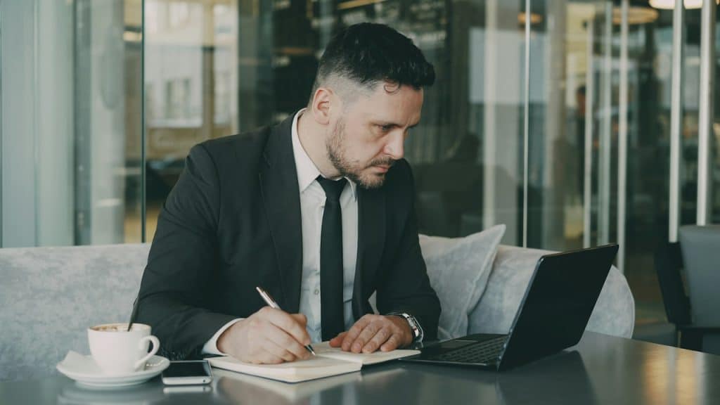 A man writing in a notebook while he looks at his laptop screen as he’s seated on a couch in his office.