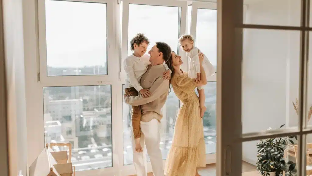 A family smiling and holding two children in a bright room with large windows.