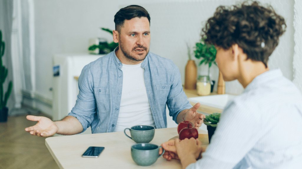 Man talks with hands open to a seated woman who is crying and covering her mouth.