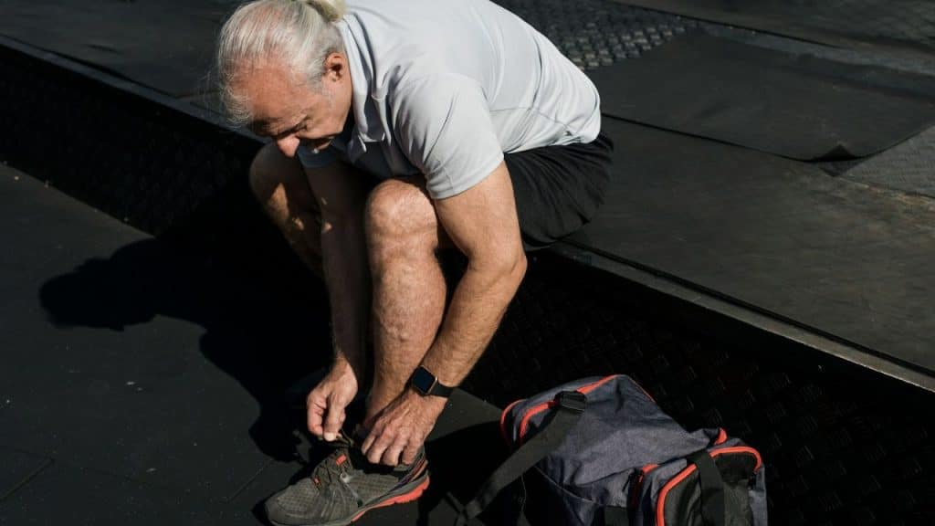 Elderly man tying shoe laces on a dark surface with a duffel bag nearby.