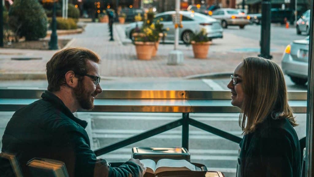 A couple sitting across from each other at a table in a cafe, looking at a book and smiling.