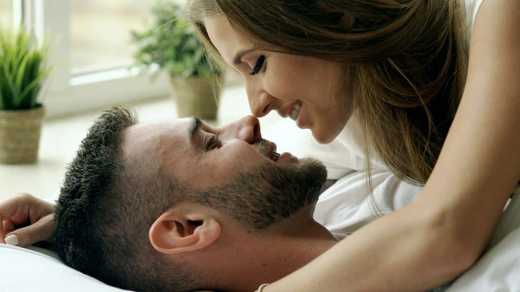 A man and woman are lying face-to-face on a bed, smiling and looking lovingly at each other.