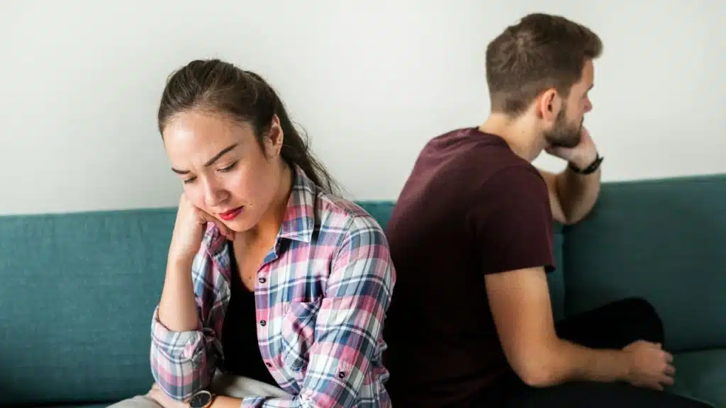 A woman sits on a teal sofa with her head down, while a man faces away.