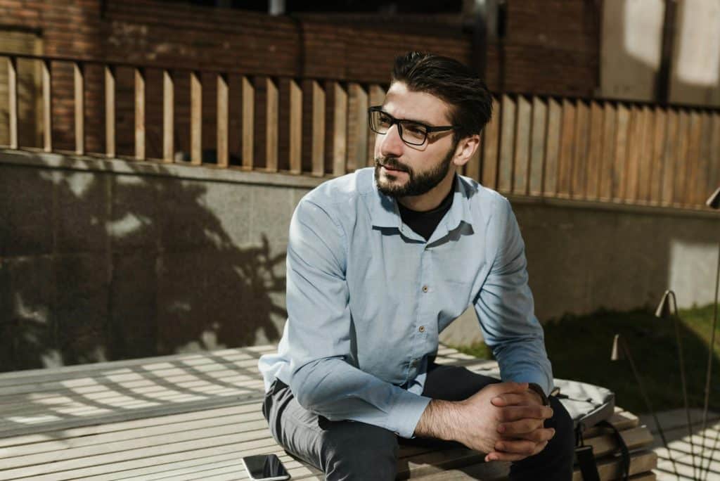 A man sits outdoors on a bench with his hands clasped, looking calmly into the distance.