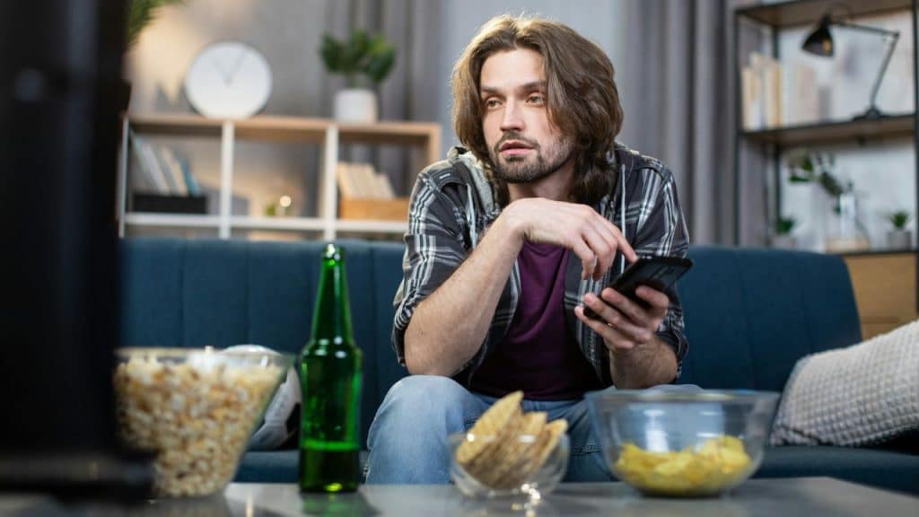 A long-haired man sits on a couch, holding a phone, with snacks and a beer nearby, watching TV.