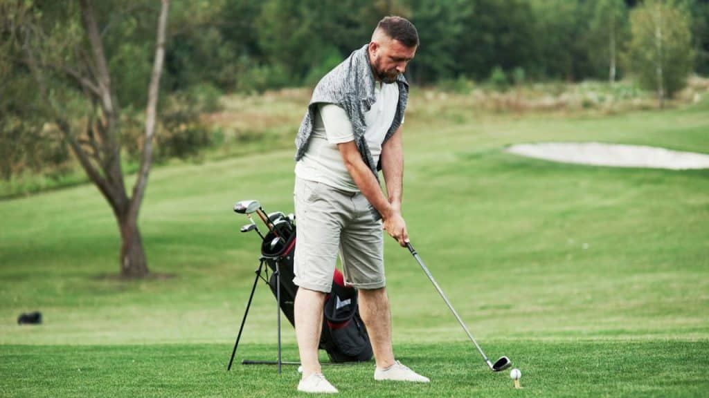 A man in golf attire swings a club toward a ball on a tee outdoors on a course.