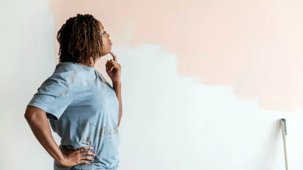 A woman in a paint-stained shirt thinks while looking at a half-painted wall.